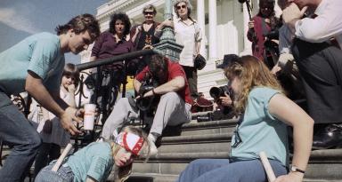 Jennifer Keelan-Chaffins wears a bandana as she crawls up the U.S. Capitol steps on her hands and knees. On her left is a man bending over and on her right a woman moves up the stairs on her back. All three protesters wear matching light blue shirts from the ADAPT organization. In front of them are media personnel with cameras and microphones recording the protest. ​​​​​[Photo Credit: Associated Press]